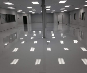 Large, empty industrial room with shiny, polished gray floors featuring USDA approved flooring. White walls and overhead fluorescent lighting reflect on the floor. Several doors, windows, and some equipment are visible in the background. Large, empty industrial room with shiny, polished gray floors featuring USDA approved flooring. White walls and overhead fluorescent lighting reflect on the floor. Several doors, windows, and some equipment are visible in the background.