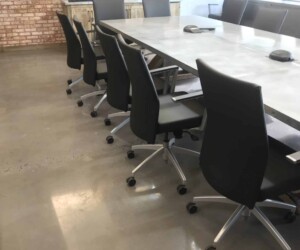 A modern conference room with a long table, several black office chairs on wheels, a large TV mounted on a brick wall, windows letting in natural light, and industrial floor coating for enhanced durability and style. A modern conference room with a long table, several black office chairs on wheels, a large TV mounted on a brick wall, windows letting in natural light, and industrial floor coating for enhanced durability and style.