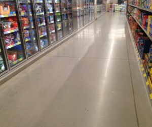 A grocery store aisle with freezers stocked with food on the left and shelves filled with packaged goods on the right; the shiny USDA approved flooring is spotless and empty. A grocery store aisle with freezers stocked with food on the left and shelves filled with packaged goods on the right; the shiny USDA approved flooring is spotless and empty.