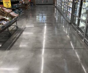 A grocery store aisle with shiny resinous flooring CT, refrigerated cases on the right, shelves with snacks on the left, and a yellow organic wheat sign near the front. The store appears clean and well-lit. A grocery store aisle with shiny resinous flooring CT, refrigerated cases on the right, shelves with snacks on the left, and a yellow organic wheat sign near the front. The store appears clean and well-lit.
