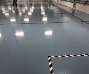 A shiny, light blue epoxy flooring Connecticut enhances this spacious indoor area, featuring black lines and a black-and-white striped border marking a section. A person is crouching in the background near the wall. A shiny, light blue epoxy flooring Connecticut enhances this spacious indoor area, featuring black lines and a black-and-white striped border marking a section. A person is crouching in the background near the wall.