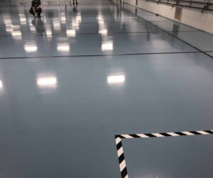 A shiny, light blue epoxy flooring Connecticut enhances this spacious indoor area, featuring black lines and a black-and-white striped border marking a section. A person is crouching in the background near the wall. A shiny, light blue epoxy flooring Connecticut enhances this spacious indoor area, featuring black lines and a black-and-white striped border marking a section. A person is crouching in the background near the wall.