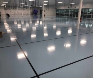 A large, empty industrial room features a shiny, polished light blue USDA approved flooring marked with black lines. Two people stand near cleaning equipment and a covered object, while bright overhead lights reflect on the pristine surface. A large, empty industrial room features a shiny, polished light blue USDA approved flooring marked with black lines. Two people stand near cleaning equipment and a covered object, while bright overhead lights reflect on the pristine surface.