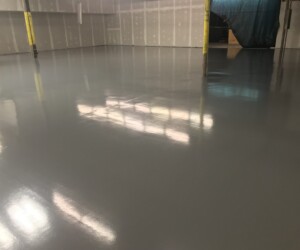 A large indoor space with freshly installed, shiny gray epoxy flooring; unfinished walls with exposed drywall and pillars are visible, and bright lights reflect off the smooth surface—perfect for those seeking premium epoxy flooring Connecticut. A large indoor space with freshly installed, shiny gray epoxy flooring; unfinished walls with exposed drywall and pillars are visible, and bright lights reflect off the smooth surface—perfect for those seeking premium epoxy flooring Connecticut.