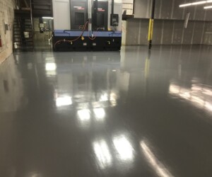 A large, empty industrial room with shiny, USDA approved flooring. In the background, a large machine with cables attached and a metal staircase leads to an upper level. Walls are unfinished with exposed material. A large, empty industrial room with shiny, USDA approved flooring. In the background, a large machine with cables attached and a metal staircase leads to an upper level. Walls are unfinished with exposed material.