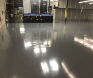 A large, empty industrial room with shiny, USDA approved flooring. In the background, a large machine with cables attached and a metal staircase leads to an upper level. Walls are unfinished with exposed material. A large, empty industrial room with shiny, USDA approved flooring. In the background, a large machine with cables attached and a metal staircase leads to an upper level. Walls are unfinished with exposed material.