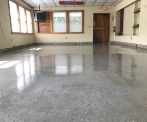 A room with anti-slip epoxy floor, beige walls, windows on the left, a mounted TV, a Budweiser sign, and a wooden door at the back. The room appears empty and well-lit. A room with anti-slip epoxy floor, beige walls, windows on the left, a mounted TV, a Budweiser sign, and a wooden door at the back. The room appears empty and well-lit.