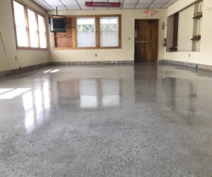 A room with anti-slip epoxy floor, beige walls, windows on the left, a mounted TV, a Budweiser sign, and a wooden door at the back. The room appears empty and well-lit. A room with anti-slip epoxy floor, beige walls, windows on the left, a mounted TV, a Budweiser sign, and a wooden door at the back. The room appears empty and well-lit.