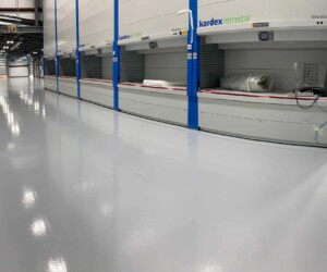 A clean, shiny, and empty industrial warehouse floor with anti-slip epoxy floor coating features automated vertical storage units labeled Kardex Remstar along the right wall; some units contain large rolled materials. A clean, shiny, and empty industrial warehouse floor with anti-slip epoxy floor coating features automated vertical storage units labeled Kardex Remstar along the right wall; some units contain large rolled materials.
