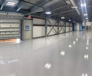 A wide, empty industrial hallway with shiny commercial kitchen flooring, exposed ceiling pipes, metal beams, overhead lights, and a partially open garage door on the left. Walls have signs and the space appears newly renovated. A wide, empty industrial hallway with shiny commercial kitchen flooring, exposed ceiling pipes, metal beams, overhead lights, and a partially open garage door on the left. Walls have signs and the space appears newly renovated.