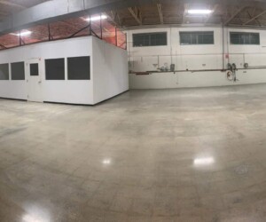 Wide-angle view of an empty industrial warehouse with polished industrial floor coating, exposed ceiling pipes, and a white enclosed office structure in the center. The space is brightly lit with overhead lights. Wide-angle view of an empty industrial warehouse with polished industrial floor coating, exposed ceiling pipes, and a white enclosed office structure in the center. The space is brightly lit with overhead lights.