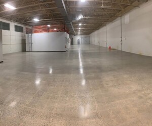 Wide view of an empty industrial warehouse with polished concrete floors and exposed ceiling beams, featuring two large white structures, a red bucket, equipment against the walls, and bright overhead lighting—ideal for epoxy flooring Connecticut projects. Wide view of an empty industrial warehouse with polished concrete floors and exposed ceiling beams, featuring two large white structures, a red bucket, equipment against the walls, and bright overhead lighting—ideal for epoxy flooring Connecticut projects.