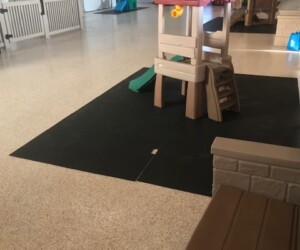 Indoor play area with plastic play structures on a black mat, including a small slide and climbing steps. The room features light-colored USDA approved flooring, a gated area, and large doors in the background. Indoor play area with plastic play structures on a black mat, including a small slide and climbing steps. The room features light-colored USDA approved flooring, a gated area, and large doors in the background.