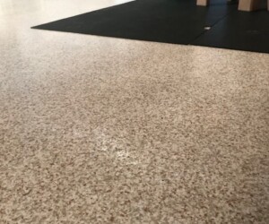 A speckled beige anti-slip epoxy floor with a black mat, part of a small green slide, and a tan play structure. In the background are radiators, a trash can, and play equipment, suggesting an indoor play area. A speckled beige anti-slip epoxy floor with a black mat, part of a small green slide, and a tan play structure. In the background are radiators, a trash can, and play equipment, suggesting an indoor play area.