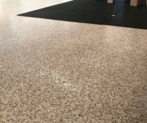 A speckled beige anti-slip epoxy floor with a black mat, part of a small green slide, and a tan play structure. In the background are radiators, a trash can, and play equipment, suggesting an indoor play area. A speckled beige anti-slip epoxy floor with a black mat, part of a small green slide, and a tan play structure. In the background are radiators, a trash can, and play equipment, suggesting an indoor play area.