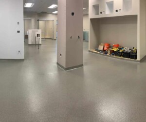 A large, empty indoor space with gray industrial floor coating and light-colored walls; shelves in the back hold construction tools and equipment, suggesting the area is under renovation. Ceiling lights illuminate the room. A large, empty indoor space with gray industrial floor coating and light-colored walls; shelves in the back hold construction tools and equipment, suggesting the area is under renovation. Ceiling lights illuminate the room.
