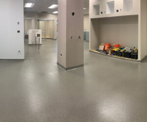 A large, empty indoor space with gray industrial floor coating and light-colored walls; shelves in the back hold construction tools and equipment, suggesting the area is under renovation. Ceiling lights illuminate the room. A large, empty indoor space with gray industrial floor coating and light-colored walls; shelves in the back hold construction tools and equipment, suggesting the area is under renovation. Ceiling lights illuminate the room.