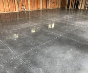 A large, smooth, and shiny industrial floor coating with visible square grid lines lies inside a building under construction; unfinished wooden framing and exposed pipes are visible along the walls. A large, smooth, and shiny industrial floor coating with visible square grid lines lies inside a building under construction; unfinished wooden framing and exposed pipes are visible along the walls.