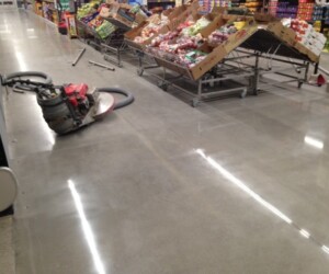 A floor buffer machine sits on a shiny, polished supermarket floor with USDA approved flooring near produce displays with fruits and vegetables in cardboard boxes. Some metal rods or pipes lie on the floor nearby. A floor buffer machine sits on a shiny, polished supermarket floor with USDA approved flooring near produce displays with fruits and vegetables in cardboard boxes. Some metal rods or pipes lie on the floor nearby.
