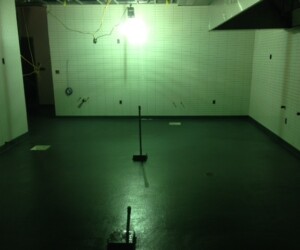 A dimly lit, empty room with white tiled walls and a dark floor features exposed pipes, wires, and a bright overhead light casting an eerie greenish tint. Two upright metal poles stand on durable industrial floor coating. A dimly lit, empty room with white tiled walls and a dark floor features exposed pipes, wires, and a bright overhead light casting an eerie greenish tint. Two upright metal poles stand on durable industrial floor coating.