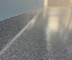Close-up view of a shiny, polished gray speckled anti-slip epoxy floor and wall base in a hallway with blurred background and overhead lights, showcasing quality epoxy flooring Connecticut installations. Close-up view of a shiny, polished gray speckled anti-slip epoxy floor and wall base in a hallway with blurred background and overhead lights, showcasing quality epoxy flooring Connecticut installations.