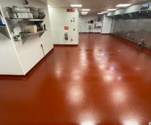 A clean, empty commercial kitchen with shiny red epoxy flooring Connecticut, stainless steel shelves and equipment lining the walls, bright overhead lights, and visible fire safety equipment. A clean, empty commercial kitchen with shiny red epoxy flooring Connecticut, stainless steel shelves and equipment lining the walls, bright overhead lights, and visible fire safety equipment.