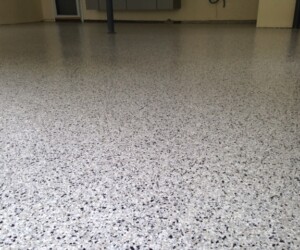 A close-up view of a smooth, speckled resinous flooring CT epoxy-coated garage floor in a mostly empty room with light-colored walls and a few fixtures in the background. A close-up view of a smooth, speckled resinous flooring CT epoxy-coated garage floor in a mostly empty room with light-colored walls and a few fixtures in the background.