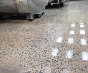 A polished concrete floor with a speckled pattern reflects overhead lights in an indoor industrial setting; anti-slip epoxy floor ensures safety, while machinery covered with protective sheets is visible in the background. A polished concrete floor with a speckled pattern reflects overhead lights in an indoor industrial setting; anti-slip epoxy floor ensures safety, while machinery covered with protective sheets is visible in the background.