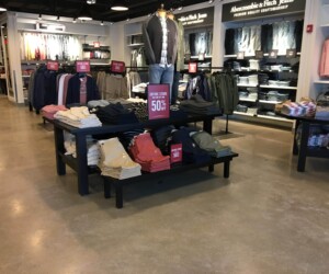 Clothing store interior with folded pants on tables, shirts hanging on racks, and a mannequin dressed in casual wear. Shelves filled with jeans and shirts line the walls, all set atop durable epoxy flooring Connecticut for a modern touch. Signs display Entire Store 50% Off. Clothing store interior with folded pants on tables, shirts hanging on racks, and a mannequin dressed in casual wear. Shelves filled with jeans and shirts line the walls, all set atop durable epoxy flooring Connecticut for a modern touch. Signs display Entire Store 50% Off.