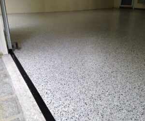 Smooth, light gray epoxy garage floor with black and white speckles, bordered by a black strip near the entryway and adjacent to a stone threshold and unfinished wall—showcasing the quality of epoxy flooring Connecticut residents trust. Smooth, light gray epoxy garage floor with black and white speckles, bordered by a black strip near the entryway and adjacent to a stone threshold and unfinished wall—showcasing the quality of epoxy flooring Connecticut residents trust.