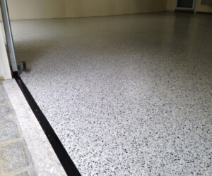 Smooth, light gray epoxy garage floor with black and white speckles, bordered by a black strip near the entryway and adjacent to a stone threshold and unfinished wall—showcasing the quality of epoxy flooring Connecticut residents trust. Smooth, light gray epoxy garage floor with black and white speckles, bordered by a black strip near the entryway and adjacent to a stone threshold and unfinished wall—showcasing the quality of epoxy flooring Connecticut residents trust.
