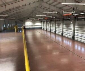 Large, empty industrial warehouse with bright overhead lights, a reddish-brown USDA approved flooring finish, a high white ceiling, multiple overhead doors on one side, and a yellow railing along an elevated walkway. Large, empty industrial warehouse with bright overhead lights, a reddish-brown USDA approved flooring finish, a high white ceiling, multiple overhead doors on one side, and a yellow railing along an elevated walkway.