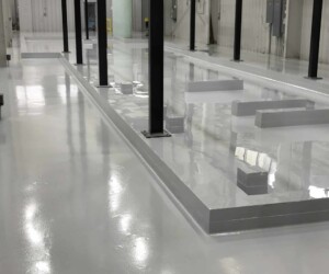 A clean, glossy, gray anti-slip epoxy floor inside an industrial facility, with black metal beams, raised platforms, and large windows along the walls. A clean, glossy, gray anti-slip epoxy floor inside an industrial facility, with black metal beams, raised platforms, and large windows along the walls.