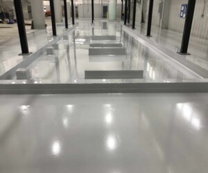 A large industrial area with a shiny, smooth grey epoxy flooring Connecticut finish. Rectangular raised sections are arranged in the center, possibly as a base for machinery. Black columns and light grey walls surround the space. A large industrial area with a shiny, smooth grey epoxy flooring Connecticut finish. Rectangular raised sections are arranged in the center, possibly as a base for machinery. Black columns and light grey walls surround the space.