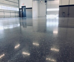A close-up view of a polished concrete floor with industrial floor coating inside an empty, well-lit garage or industrial space, with large windows and garage doors visible in the background. A close-up view of a polished concrete floor with industrial floor coating inside an empty, well-lit garage or industrial space, with large windows and garage doors visible in the background.