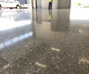 Close-up view of a polished concrete floor with industrial floor coating inside a building, reflecting light and part of a person standing in the background, with a parked vehicle visible outside through open doors. Close-up view of a polished concrete floor with industrial floor coating inside a building, reflecting light and part of a person standing in the background, with a parked vehicle visible outside through open doors.