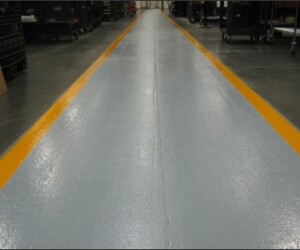 A smooth, light gray walkway with bright yellow edges runs through a warehouse or industrial facility, featuring industrial floor coating and surrounded by dark flooring and equipment. A smooth, light gray walkway with bright yellow edges runs through a warehouse or industrial facility, featuring industrial floor coating and surrounded by dark flooring and equipment.