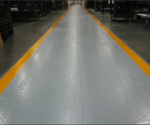 A smooth, light gray walkway with bright yellow edges runs through a warehouse or industrial facility, featuring industrial floor coating and surrounded by dark flooring and equipment. A smooth, light gray walkway with bright yellow edges runs through a warehouse or industrial facility, featuring industrial floor coating and surrounded by dark flooring and equipment.