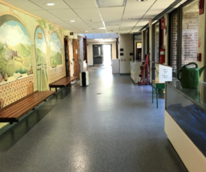 A brightly lit hallway with durable epoxy flooring Connecticut features a mural on the left wall, wooden benches, windows to the right, a water dispenser, and a green watering can. At the end, a door opens to an outdoor area. A brightly lit hallway with durable epoxy flooring Connecticut features a mural on the left wall, wooden benches, windows to the right, a water dispenser, and a green watering can. At the end, a door opens to an outdoor area.