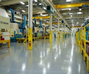Large, brightly lit factory floor with yellow safety railings, polished industrial floor coating, overhead cranes, and industrial machines. The space appears clean and organized, with equipment lining the walls. Large, brightly lit factory floor with yellow safety railings, polished industrial floor coating, overhead cranes, and industrial machines. The space appears clean and organized, with equipment lining the walls.