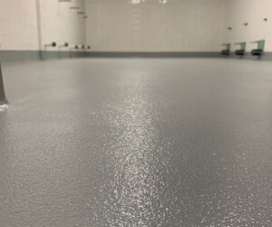 Low-angle view of an empty room with commercial kitchen flooring, featuring a textured gray anti-slip epoxy floor, white tiled walls, red and white checkered trim near the ceiling, and metal fixtures on the walls. Low-angle view of an empty room with commercial kitchen flooring, featuring a textured gray anti-slip epoxy floor, white tiled walls, red and white checkered trim near the ceiling, and metal fixtures on the walls.