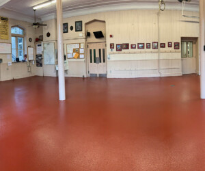 A wide, empty room with industrial floor coating, red resinous flooring CT, white walls, large windows, and garage doors. Framed photos and items hang on the walls; tables, chairs, and coffee machines sit to one side. Two white support columns stand in the center. A wide, empty room with industrial floor coating, red resinous flooring CT, white walls, large windows, and garage doors. Framed photos and items hang on the walls; tables, chairs, and coffee machines sit to one side. Two white support columns stand in the center.