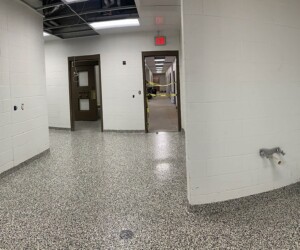 A wide, panoramic view of a clean, empty hallway with white brick walls, USDA approved flooring beneath speckled surfaces, exposed ceiling, brown doors, and some visible wires; the lighting is bright with no people present. A wide, panoramic view of a clean, empty hallway with white brick walls, USDA approved flooring beneath speckled surfaces, exposed ceiling, brown doors, and some visible wires; the lighting is bright with no people present.