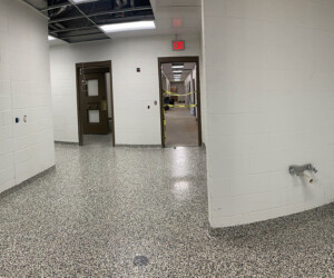 A wide, panoramic view of a clean, empty hallway with white brick walls, USDA approved flooring beneath speckled surfaces, exposed ceiling, brown doors, and some visible wires; the lighting is bright with no people present. A wide, panoramic view of a clean, empty hallway with white brick walls, USDA approved flooring beneath speckled surfaces, exposed ceiling, brown doors, and some visible wires; the lighting is bright with no people present.