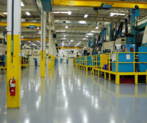 A clean, brightly lit factory floor with yellow railings, industrial machines, and overhead cranes features shiny industrial floor coating that reflects the lighting. A red fire extinguisher is mounted on a yellow pillar to the left. A clean, brightly lit factory floor with yellow railings, industrial machines, and overhead cranes features shiny industrial floor coating that reflects the lighting. A red fire extinguisher is mounted on a yellow pillar to the left.
