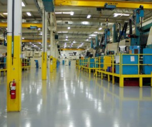 A clean, brightly lit factory floor with yellow railings, industrial machines, and overhead cranes features shiny industrial floor coating that reflects the lighting. A red fire extinguisher is mounted on a yellow pillar to the left. A clean, brightly lit factory floor with yellow railings, industrial machines, and overhead cranes features shiny industrial floor coating that reflects the lighting. A red fire extinguisher is mounted on a yellow pillar to the left.