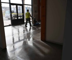 A person in a yellow shirt operates a floor cleaning machine on a shiny, polished concrete floor with industrial floor coating near large glass doors and sunlight streaming in. A truck is parked outside the building. A person in a yellow shirt operates a floor cleaning machine on a shiny, polished concrete floor with industrial floor coating near large glass doors and sunlight streaming in. A truck is parked outside the building.