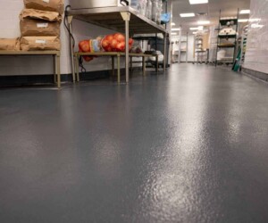 A commercial kitchen with clean, textured dark gray USDA approved flooring, metal shelving holding bags and onions, white tiled walls, and bright overhead lighting. A commercial kitchen with clean, textured dark gray USDA approved flooring, metal shelving holding bags and onions, white tiled walls, and bright overhead lighting.