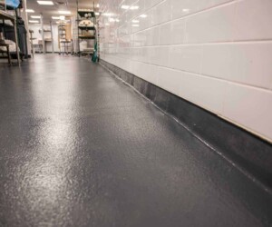 A clean, shiny gray anti-slip epoxy floor with a coved base meets a white tiled wall in a commercial kitchen or facility, with equipment and shelves visible in the background. A clean, shiny gray anti-slip epoxy floor with a coved base meets a white tiled wall in a commercial kitchen or facility, with equipment and shelves visible in the background.