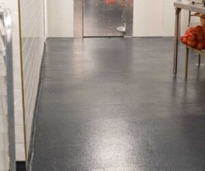 A clean commercial kitchen area with USDA approved flooring, a shiny dark surface, metal prep table holding onions on the right, white tiled wall on the left, and a closed walk-in freezer door at the end of the room. A clean commercial kitchen area with USDA approved flooring, a shiny dark surface, metal prep table holding onions on the right, white tiled wall on the left, and a closed walk-in freezer door at the end of the room.
